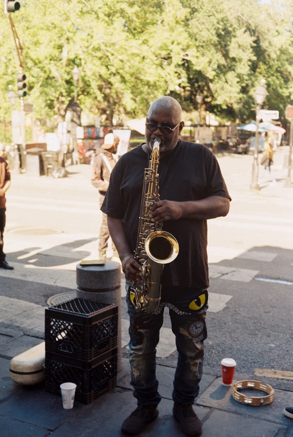 A street musician is standing in on a sidewalk and front of a cross walk with some people and trees which are in the bright sunshine. The musician standing in the shade is an older black man with short gray hair is playing a large shiny saxophone wearing a wearing t-shirt and black jeans with serval colorful patches.