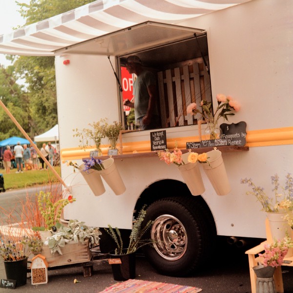 A white trailer with an open sale window with a striped awning above it, a person can be visible inside looking out the open back. The window has a ledge with cans hanging off of it holding flower stems and signs advertising the prices per stem ranging from $2 to $8. There are also pots with flowers and plants on the ground outside the trailer.